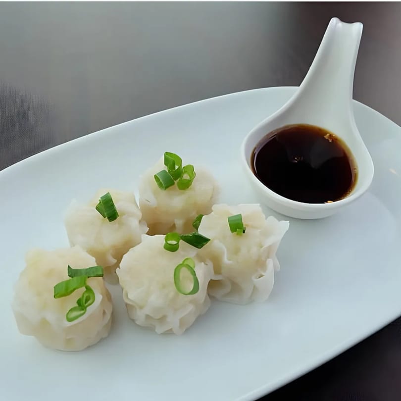 Five steamed dumplings topped with green onions, served with a side of dark sauce on a white plate.