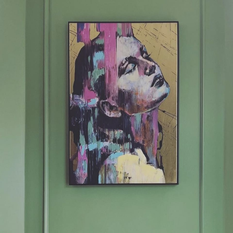 Abstract painting of a woman's face with colorful brushstrokes on a gold background, hanging on a green wall.