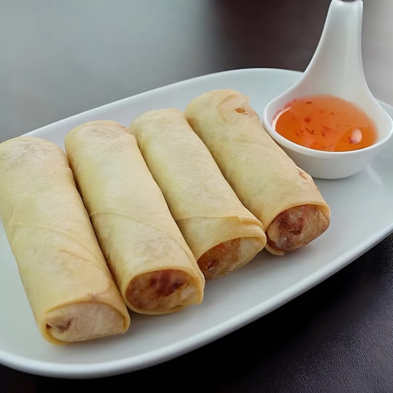 Four golden-brown Thai spring rolls with sweet chili dipping sauce on a white rectangular plate.