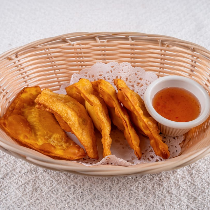 Golden fried wontons and sweet chili dipping sauce in a woven basket on a white tablecloth.
