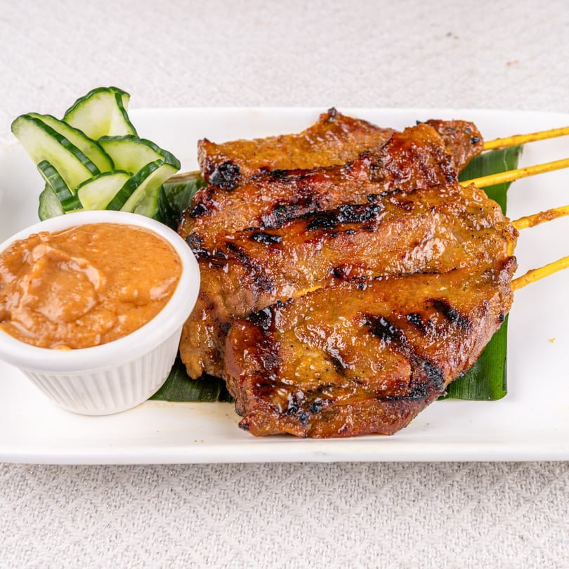 Grilled Thai chicken satay skewers with peanut sauce and cucumber slices on a white plate.