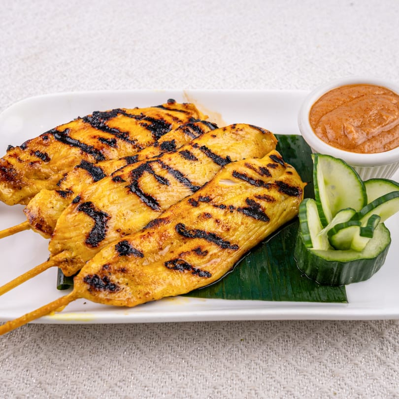 Three grilled chicken satay skewers with peanut sauce and cucumber slices on a white plate.